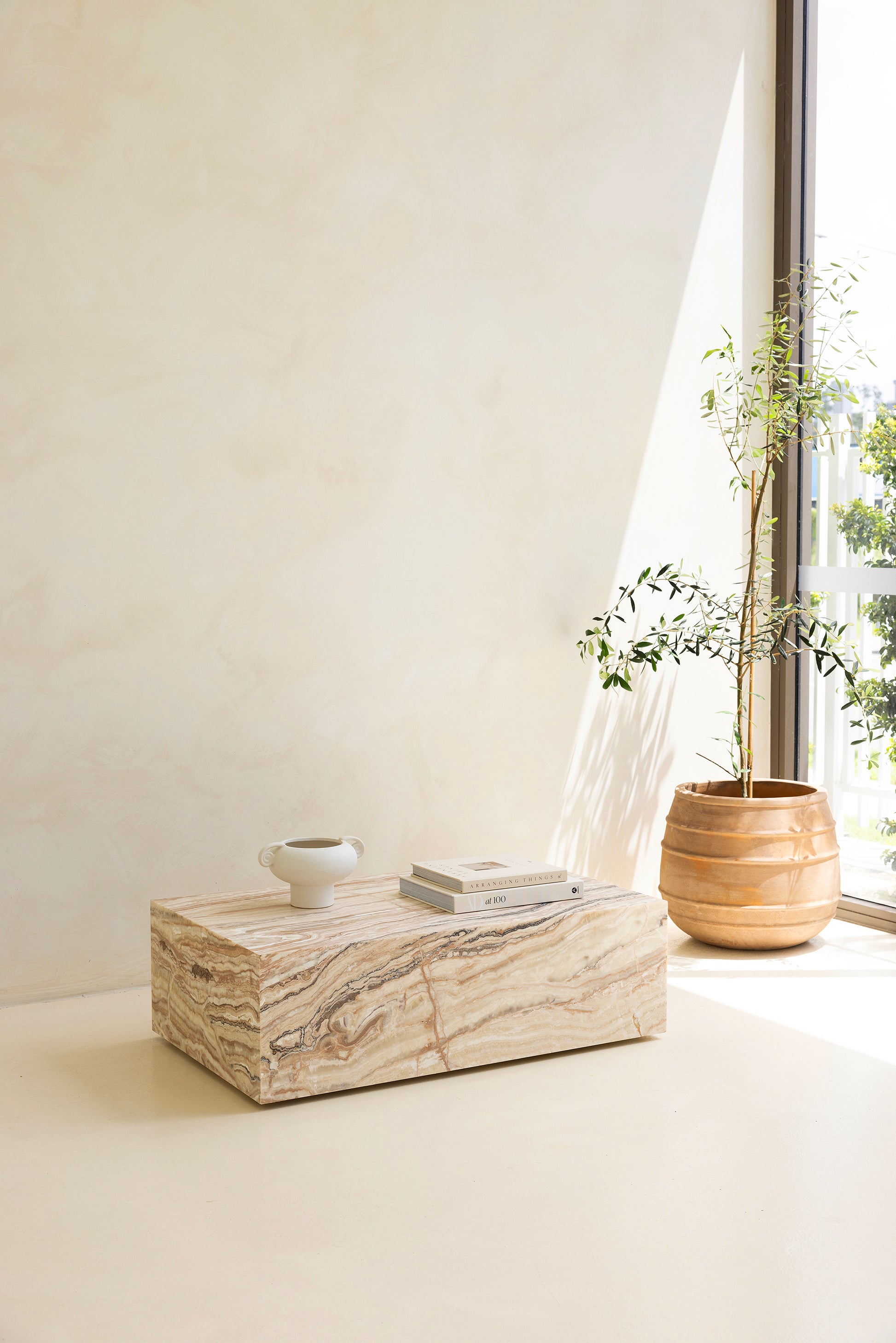 A marble coffee table with a sculptural design, placed indoors, with a plant and a wooden chair in the background.