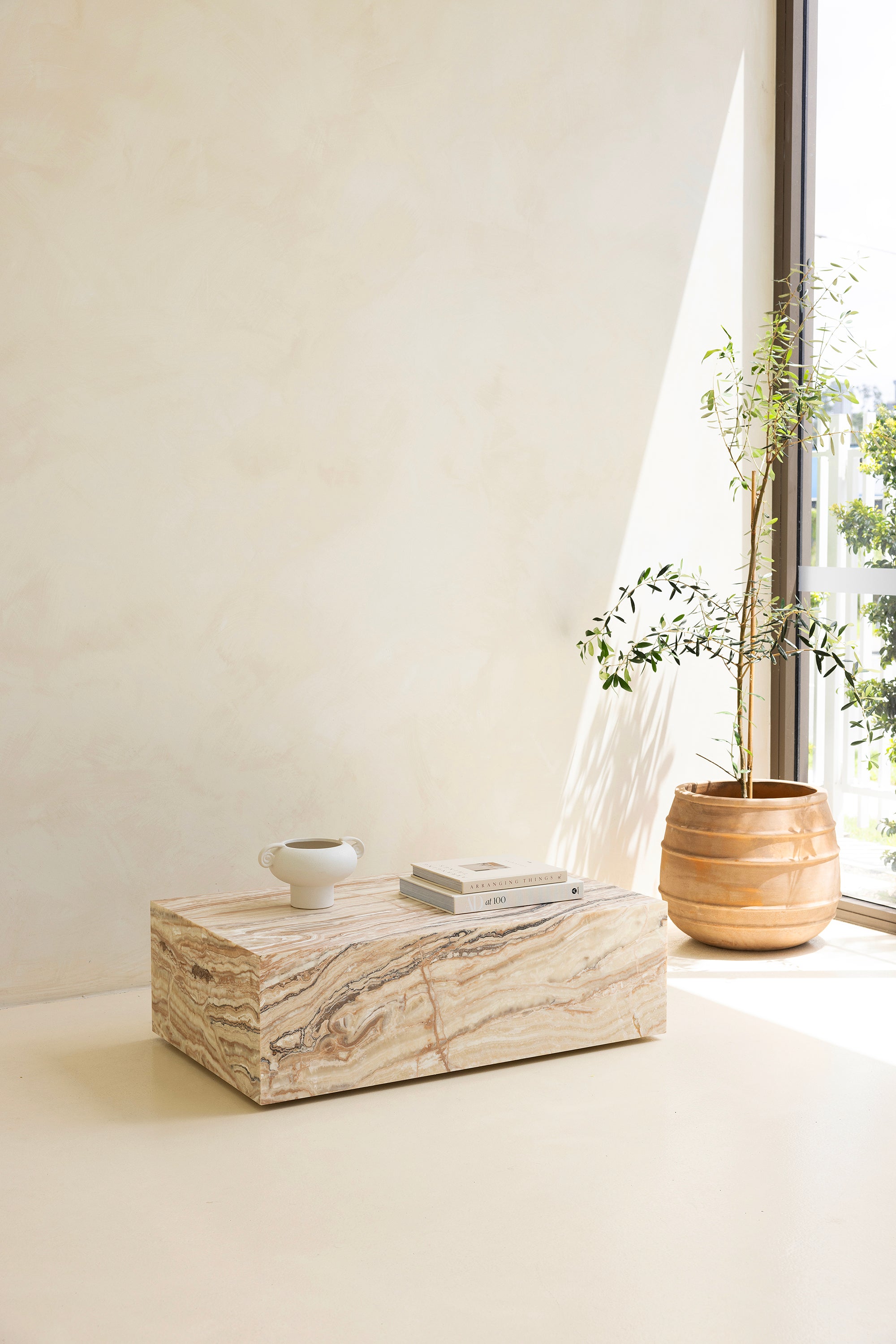 A marble coffee table with a sculptural design, placed indoors, with a plant and a wooden chair in the background.