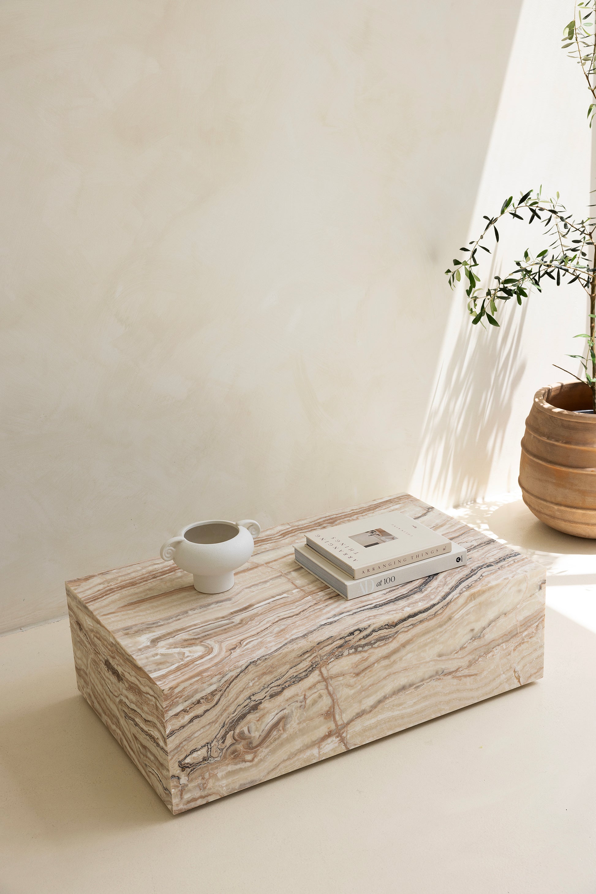 A marble coffee table with a sculptural design, placed indoors, with a plant and a wooden chair in the background.