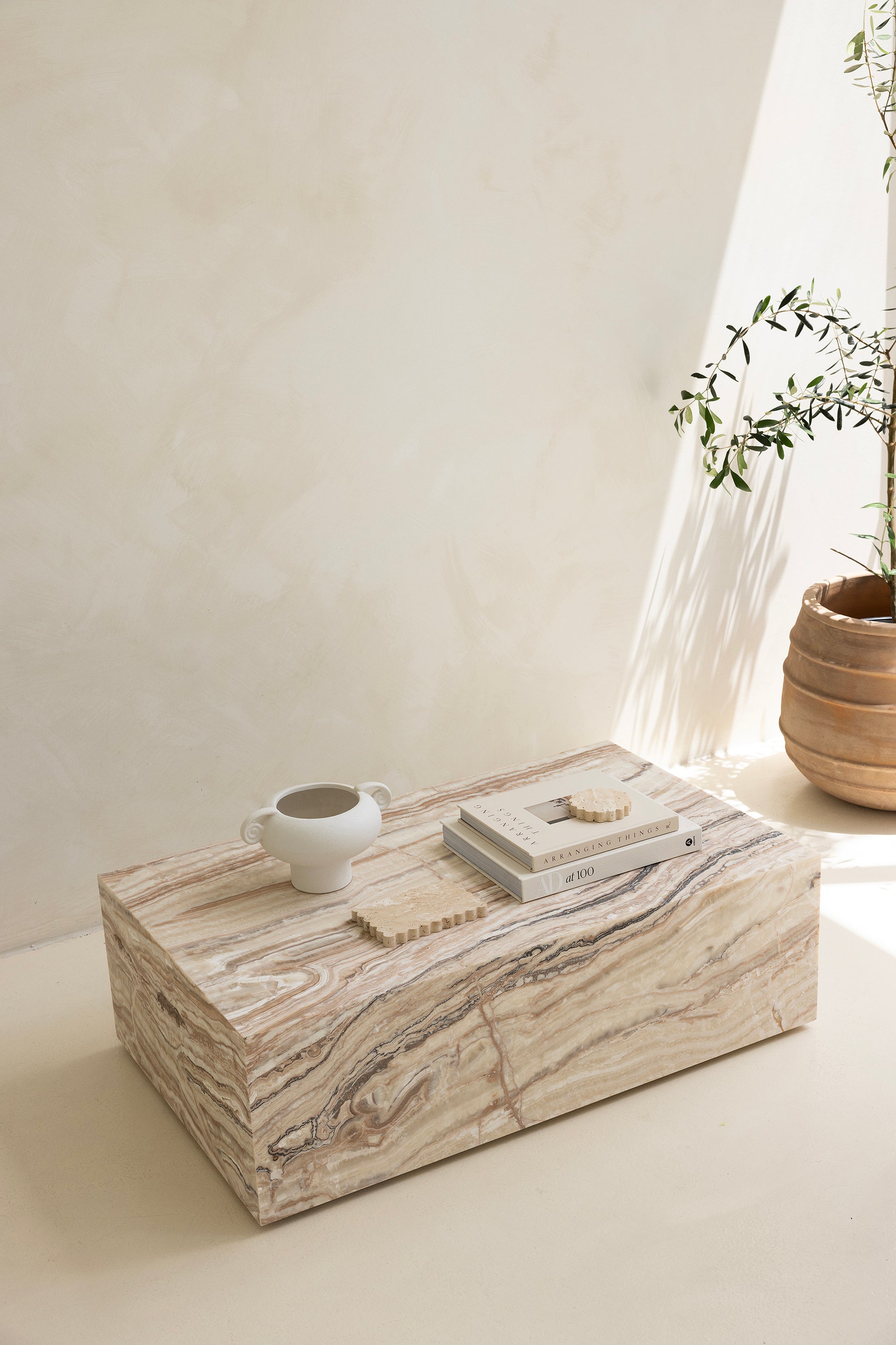 A marble coffee table with a sculptural design, placed indoors, with a plant and a wooden chair in the background.