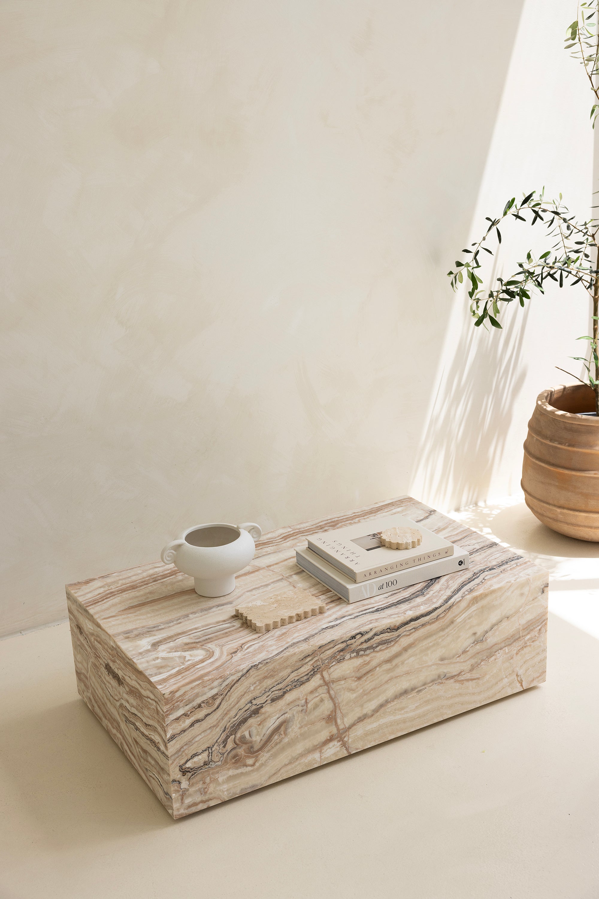 A marble coffee table with a sculptural design, placed indoors, with a plant and a wooden chair in the background.