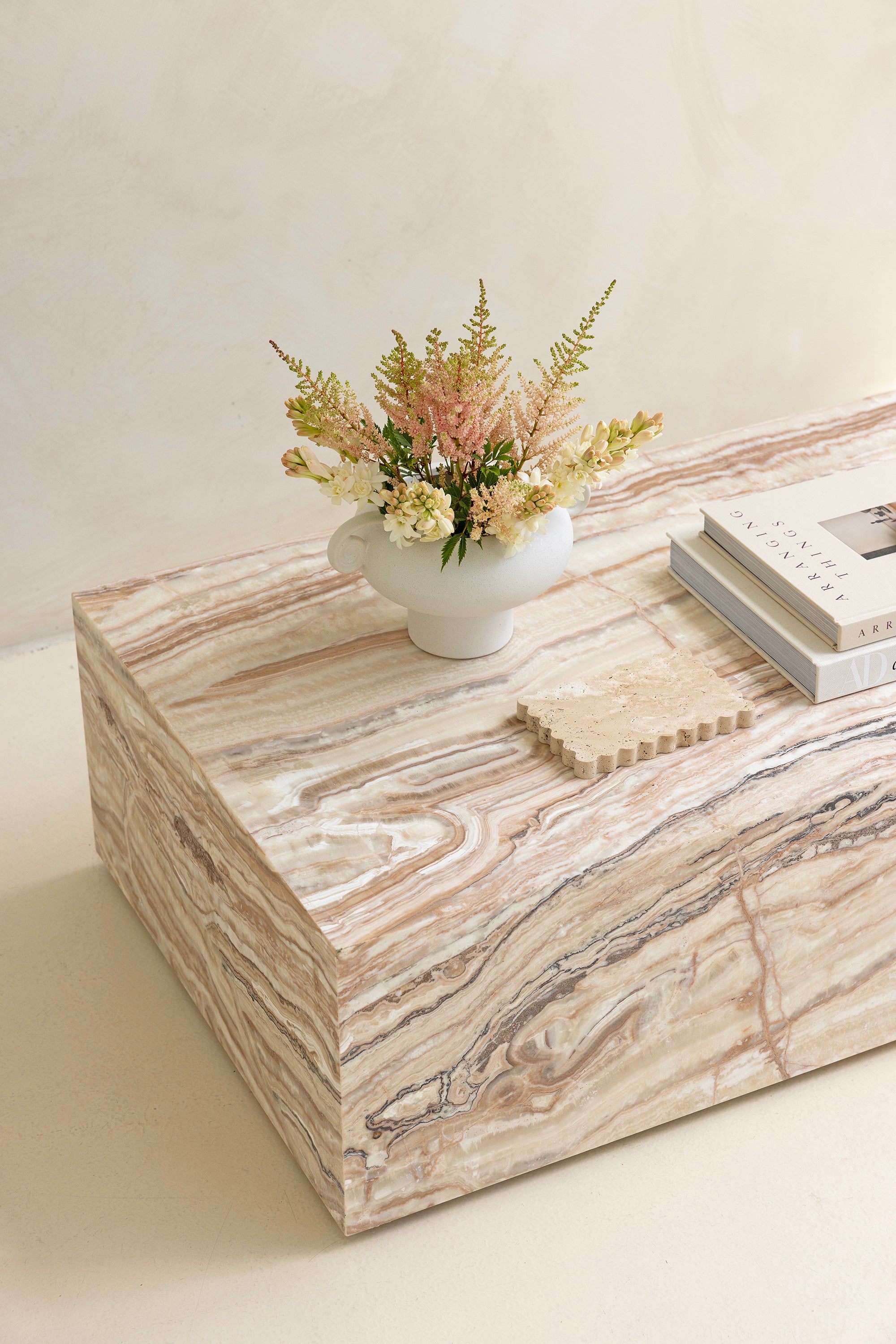 A marble coffee table with a sculptural design, placed indoors, with a plant and a wooden chair in the background.