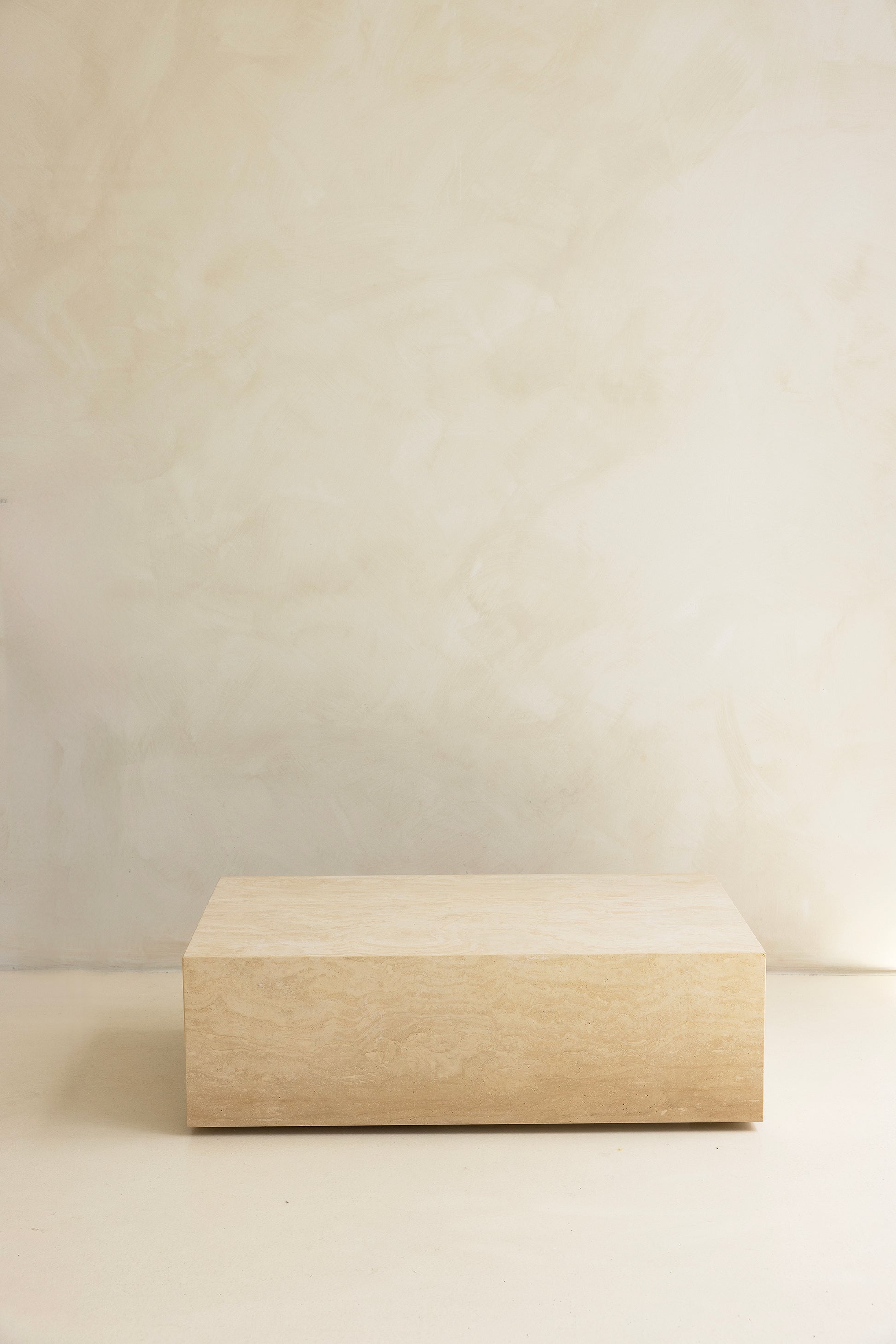 A marble coffee table with a sculptural design, placed indoors, with a plant and a wooden chair in the background.