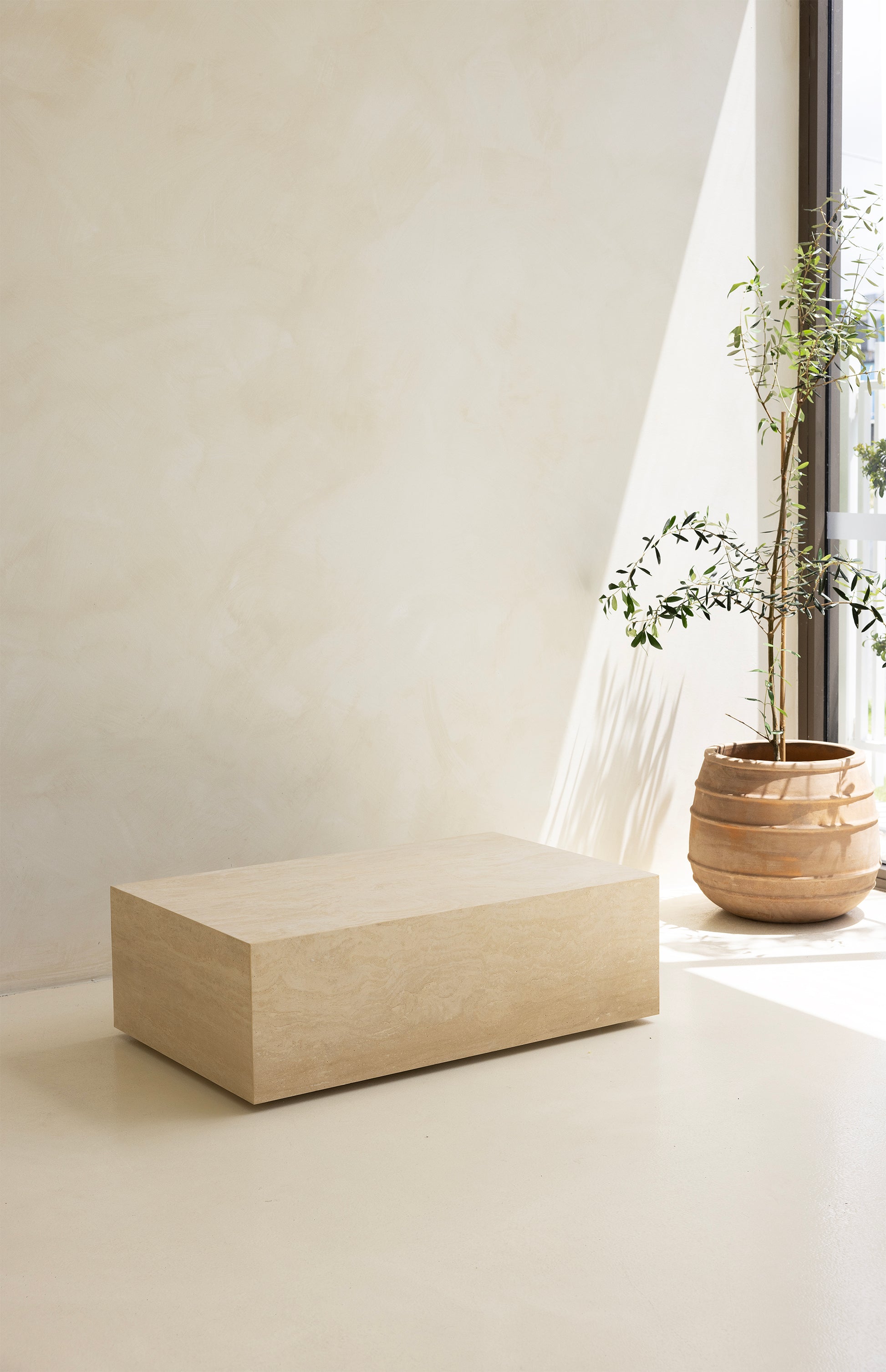 A marble coffee table with a sculptural design, placed indoors, with a plant and a wooden chair in the background.