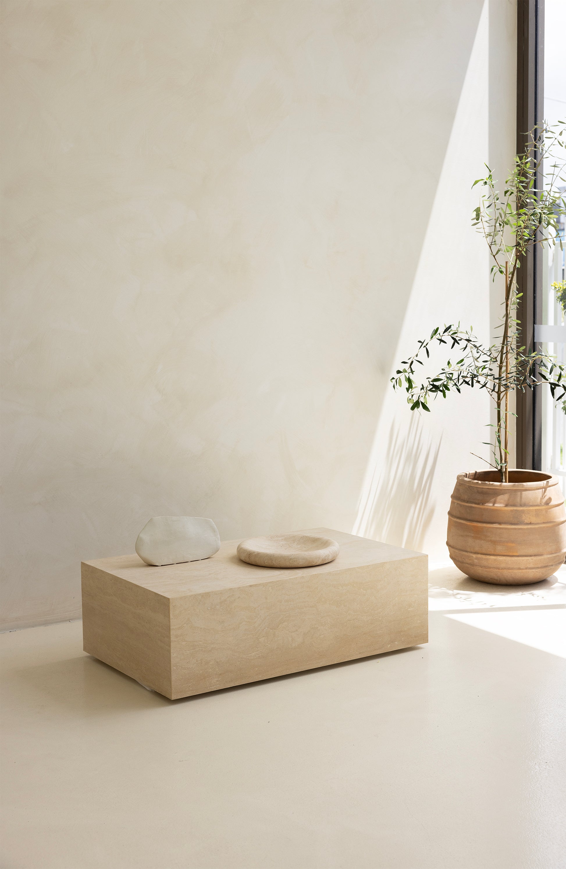 A marble coffee table with a sculptural design, placed indoors, with a plant and a wooden chair in the background.