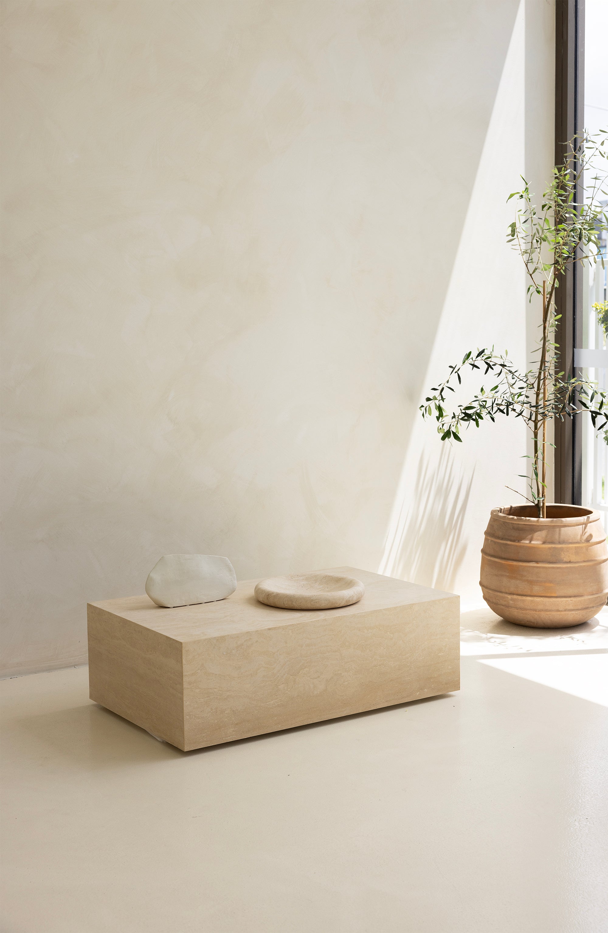 A marble coffee table with a sculptural design, placed indoors, with a plant and a wooden chair in the background.