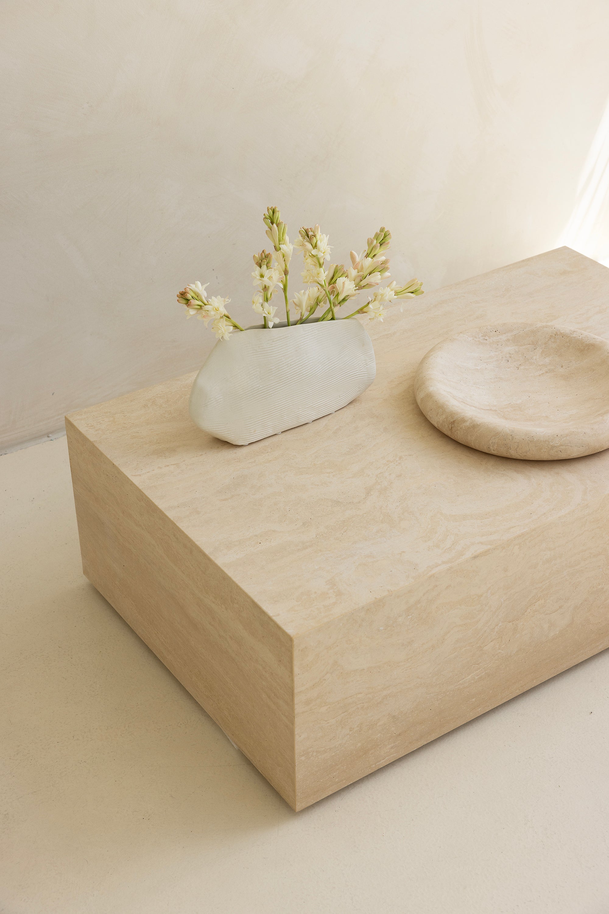 A marble coffee table with a sculptural design, placed indoors, with a plant and a wooden chair in the background.