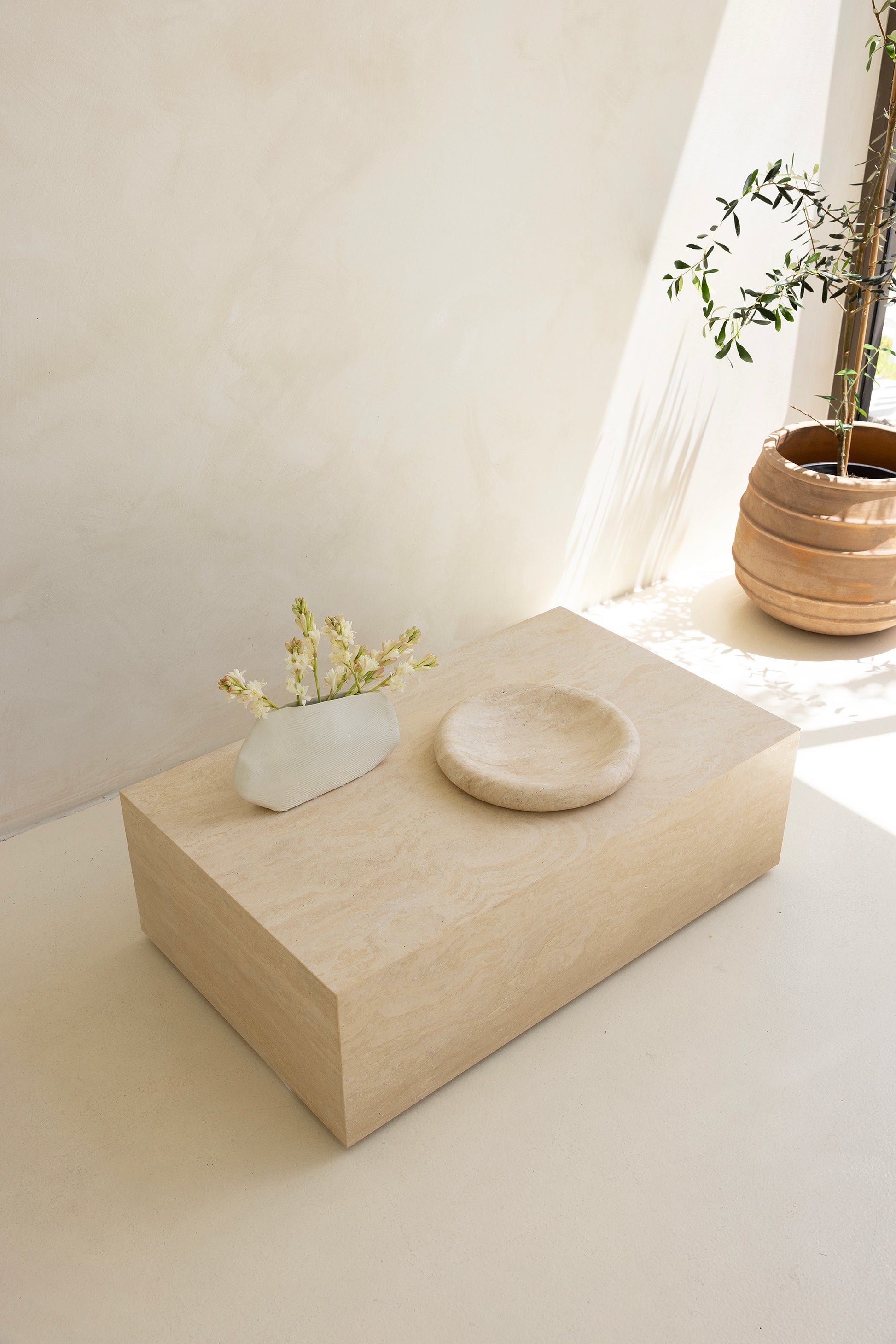 A marble coffee table with a sculptural design, placed indoors, with a plant and a wooden chair in the background.
