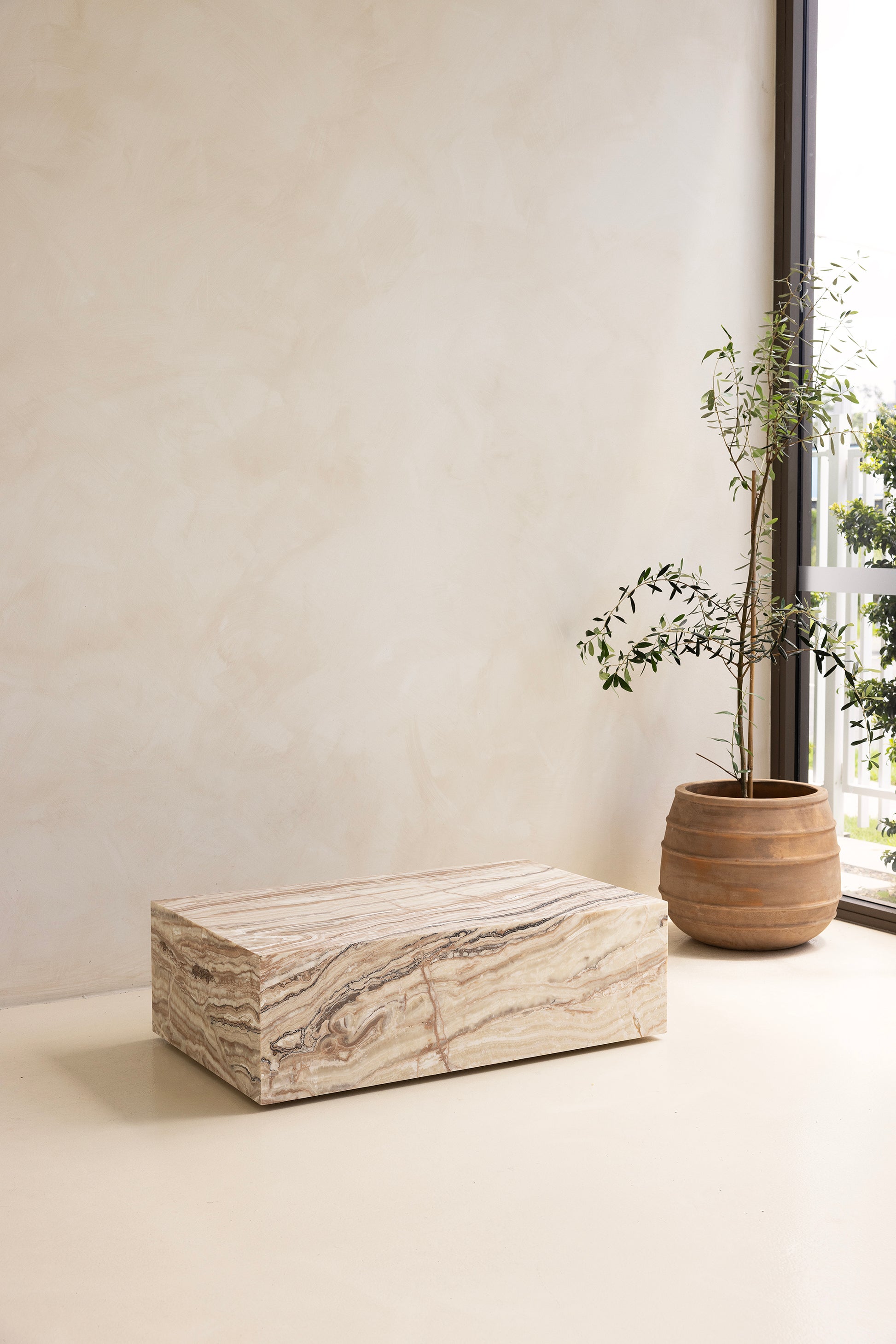 A marble coffee table with a sculptural design, placed indoors, with a plant and a wooden chair in the background.