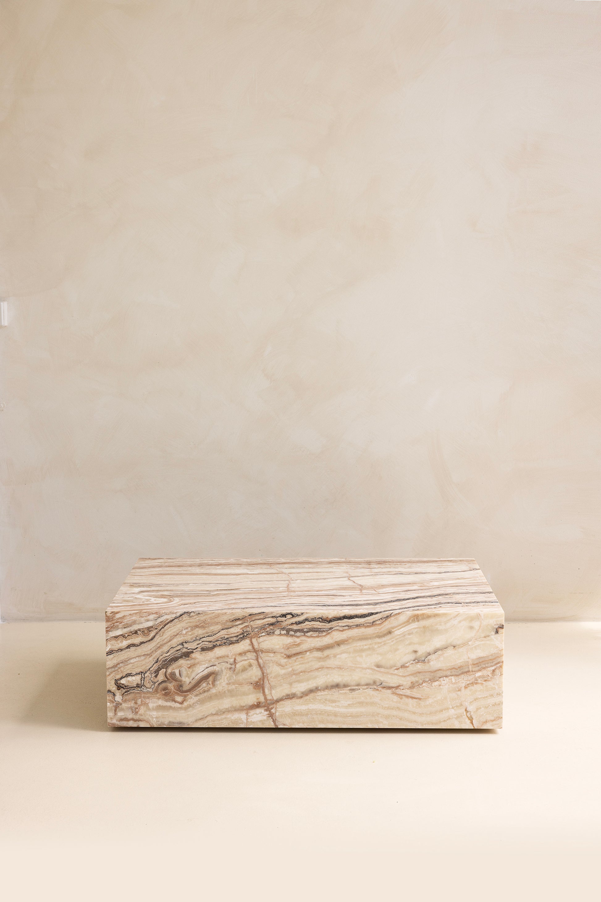A marble coffee table with a sculptural design, placed indoors, with a plant and a wooden chair in the background.