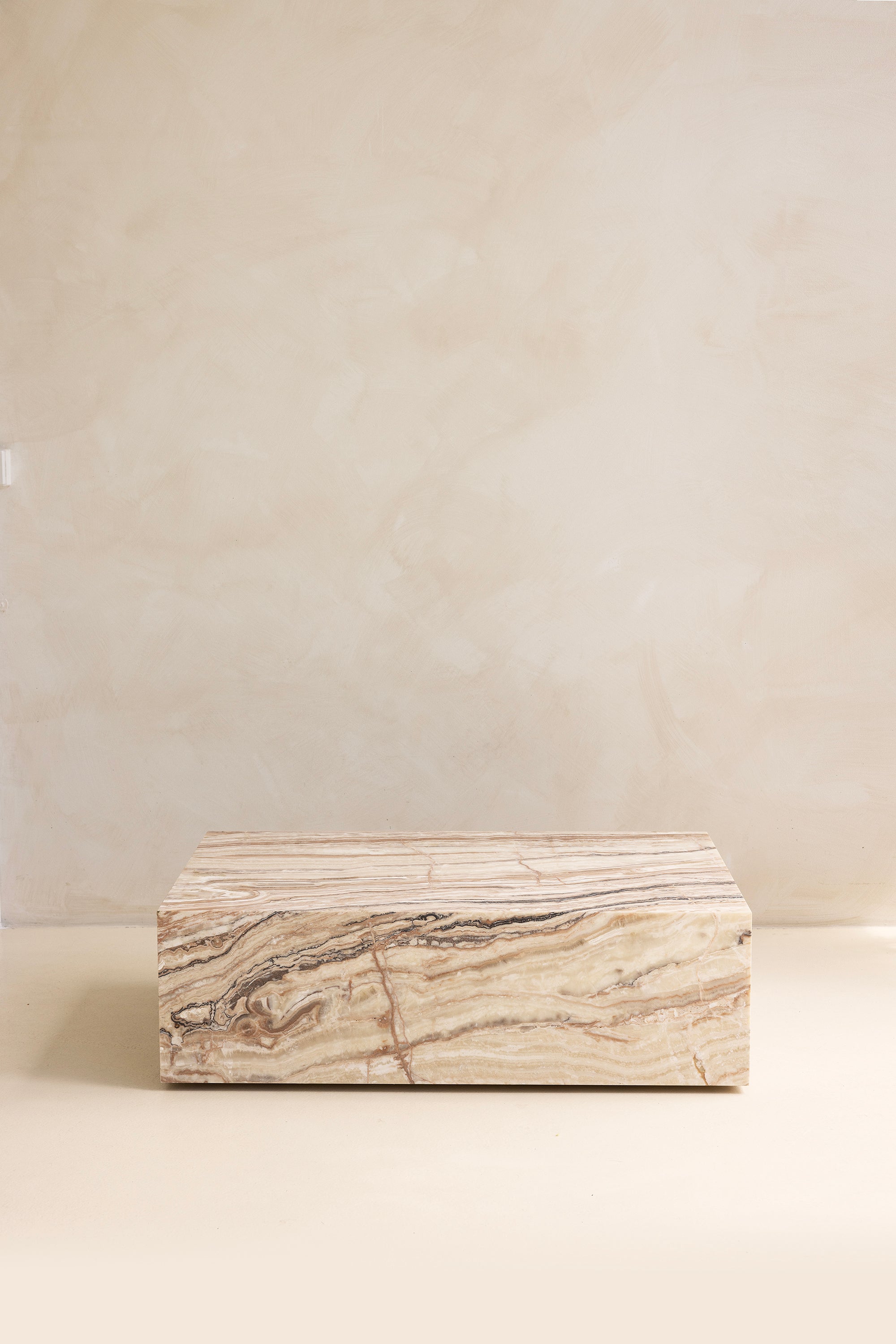 A marble coffee table with a sculptural design, placed indoors, with a plant and a wooden chair in the background.